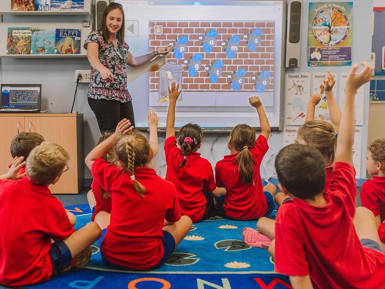 A teacher leads a Phonics Hero game on the classroom whiteboard as pupils raise their hands
